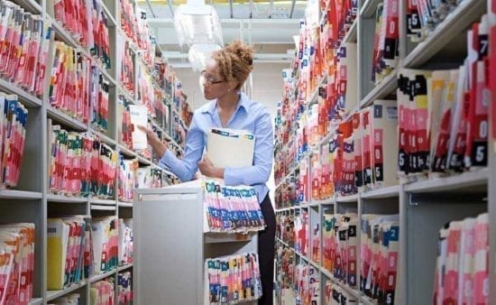a female health inforamtion technician standing between to tall shelves and filing papers
